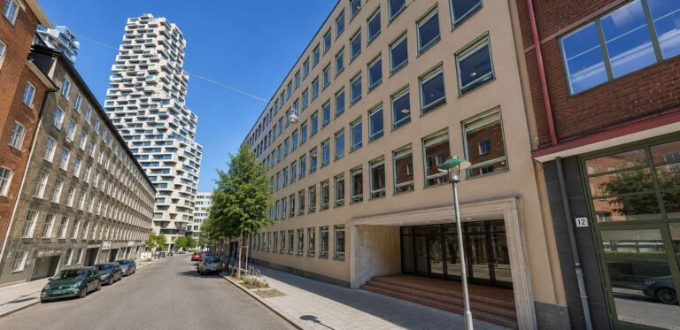 Modern kontorsbyggnad i Hagastaden med blå himmel och träd längs gatan.