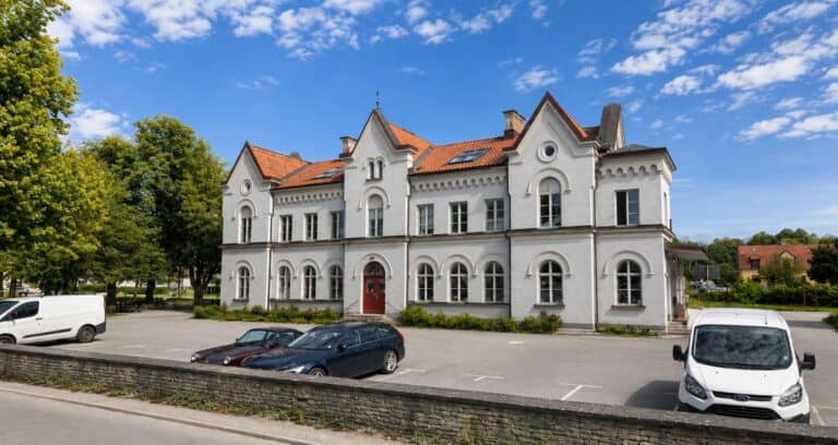 Vit gammal byggnad med röd dörr och parkering framför.