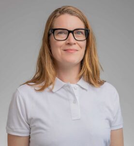 Kvinna med glasögon och vitt polo mot grå bakgrund.
