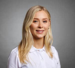 Ung kvinna med blont hår och vit skjorta ler mot kameran.