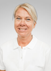 Kvinna med kort blond hår och vit polo, ler mot kameran.