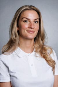 Kvinna med långt blont hår och vit polo, leende mot kameran.