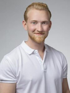 Ung man med blont hår och skägg, i vit skjorta, ler mot kameran.