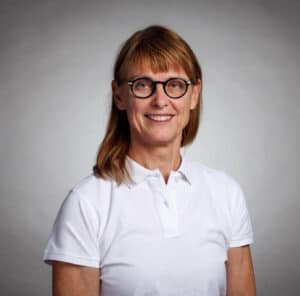 Kvinna med glasögon och vitt polo mot grå bakgrund.