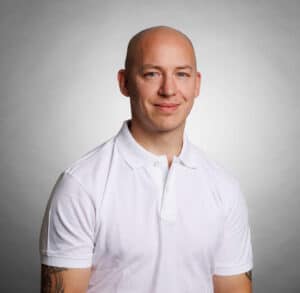 Man i vit polo shirt med tatueringar på armarna, leende mot kameran.