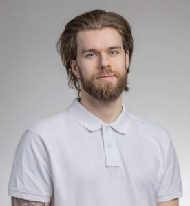 Ung man med långt hår och skägg i vit polo mot grå bakgrund.