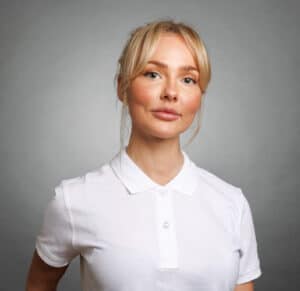 Ung kvinna med blont hår och vit polo mot grå bakgrund.
