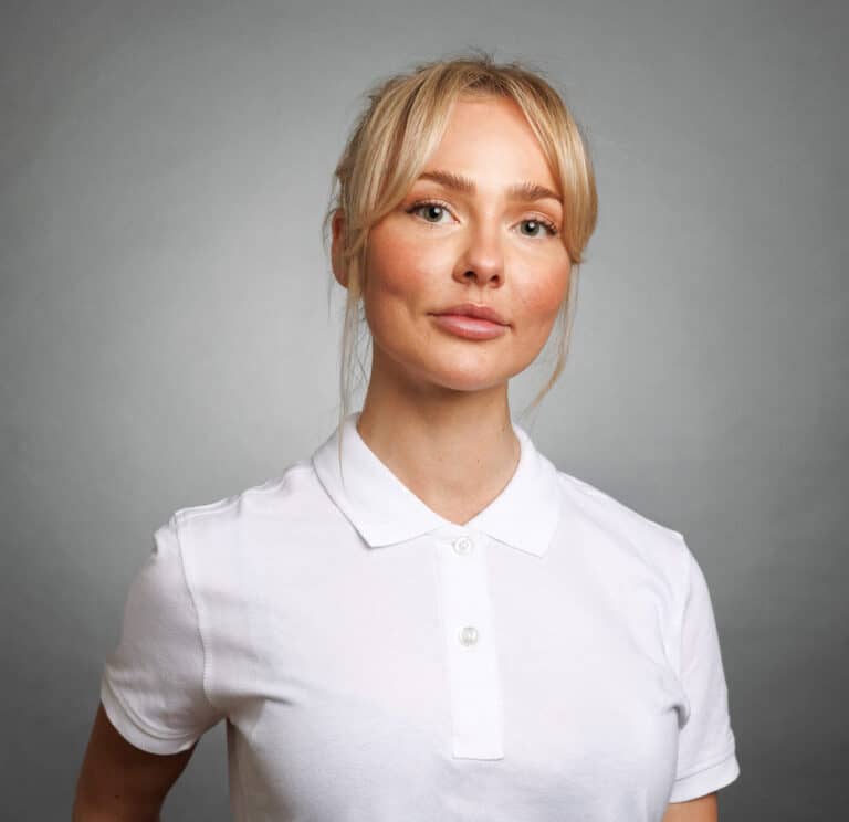 Ung kvinna med blont hår och vit polo mot grå bakgrund.