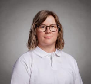 Kvinna med glasögon och vitt polo mot grå bakgrund.