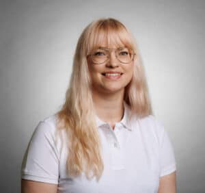 Kvinna med blont hår och glasögon, ler mot kameran.