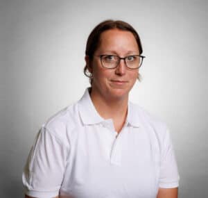 Kvinna med glasögon i vit polo mot grå bakgrund.