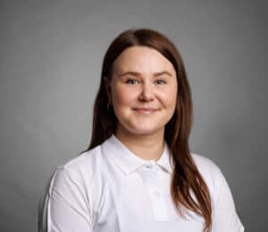 Mild smile woman with brown hair in white shirt against gray background.