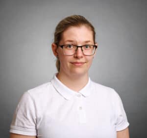 Kvinna med glasögon och vitt polo mot grå bakgrund.