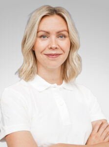 Kvinna med blont, vågigt hår och vit polo mot ljusgrå bakgrund.