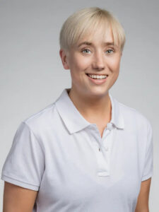 Ung kvinna med kort blont hår och vit polo-shirt ler mot kameran.