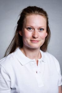 Kvinna med långt brunt hår och vit polo mot grå bakgrund.