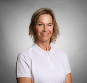 Kvinna i vit polo mot grå bakgrund, ler och tittar mot kameran.