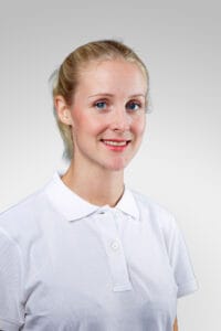 Kvinna i vit polo med blont hår och blå ögon ler mot kameran.