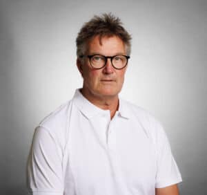 Man med glasögon och vit polo mot grå bakgrund.