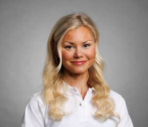 Glad kvinna med blont hår och vit skjorta mot grå bakgrund.