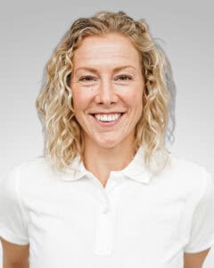Kvinna med blont, lockigt hår och vit skjorta ler mot kameran.