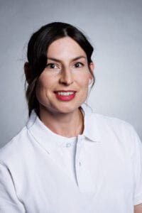 Kvinna med mörkt hår och vit skjorta, leende mot kameran.