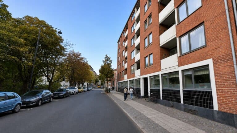 Trädkantad gata i Lund med rött flerbostadshus och lokaler i gatuplan.
