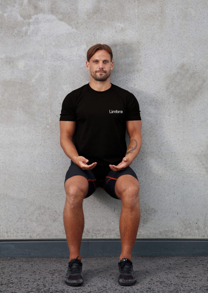 Man i svart t-shirt och shorts gör övning sittande med ryggen mot väggen